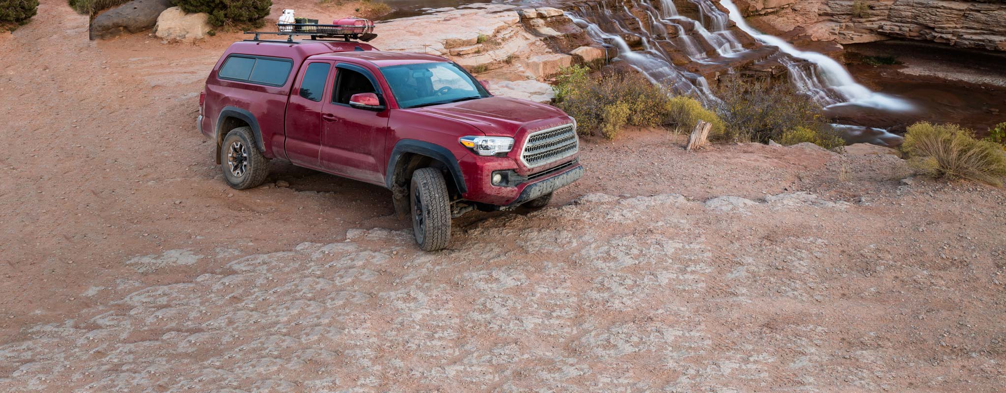 Red pickup with canopy on sandy ground near a waterfall.